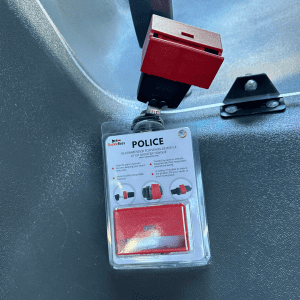 Attached to a car's interior, a clear plastic package labeled 'BuckleBoss POLICE' displays a red seatbelt guard and instructions, designed to secure passengers.