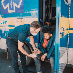 A paramedic in a dark blue uniform and face mask attends to a young man with a beard, who is seated and wrapped in a blue blanket by the open door of an ambulance