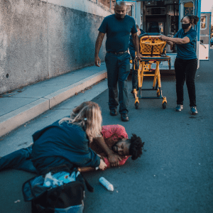 A paramedic team urgently assists a man lying on the pavement, as a bystander provides comfort.