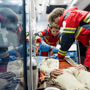 Emergency medical technicians (EMTs), a man and a woman in red and yellow reflective uniforms, provide urgent medical care to a patient inside an ambulance