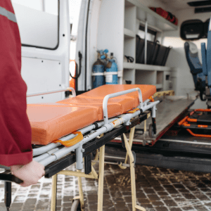 A medical professional in a red uniform is loading an orange stretcher into the back of an ambulance.