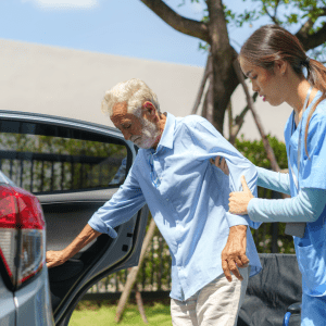 A young female caregiver in blue scrubs assists an elderly man with a white beard and blue shirt as he exits a car under a bright sunny sky.