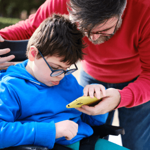 A man helps a young boy with special needs use a smartphone while the boy sits in a wheelchair.