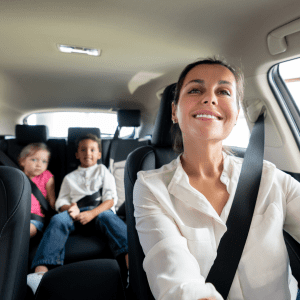 A mother in a white blouse smiles at the camera while driving, with her two children securely fastened in car seats behind her. The young girl wears a pink top, and the boy sports a white shirt.