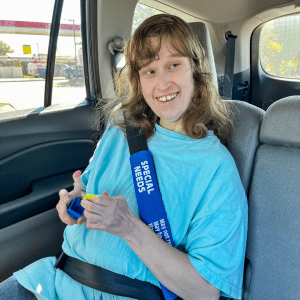 A cheerful young woman with light brown hair, wearing a light blue t-shirt that reads 'SPECIAL NEEDS,' is seated in a car with a blue 'SPECIAL NEEDS' seatbelt cover.