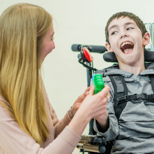 A heartwarming scene of a woman and a young boy sharing a joyful moment. The woman, with long blonde hair and wearing a pink sweater, is interacting with the boy who is seated in a wheelchair, holding a green toy.