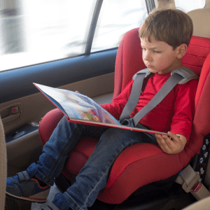 A young boy, dressed in a red shirt and blue jeans, is seated in a car booster seat, deeply focused on reading a colorful children's book.