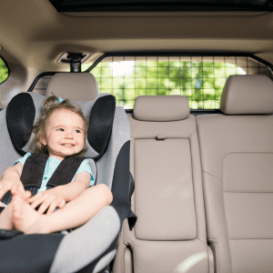 A cheerful toddler girl, with light brown hair tied up with a blue bow, smiles while securely seated in a modern gray car seat.