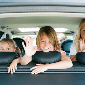 Three young children in the backseat of a car, smiling and waving at the camera during a road trip. Two girls in the front row are waving with their hands visible over the seat headrests.