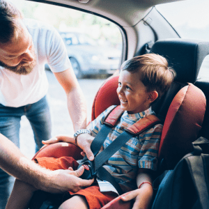 A young boy, smiling broadly, is securely fastened in a red child car seat by his father.