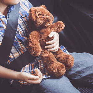 Close-up of a young child in a plaid shirt, securely fastened by a seatbelt in a car seat, holding a well-loved brown teddy bear.