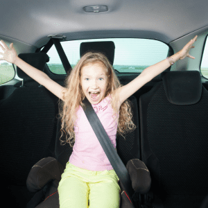 A young girl with curly blonde hair, wearing a pink shirt and lime green pants, joyfully raises her arms and exclaims in excitement while securely buckled in the backseat of a car.